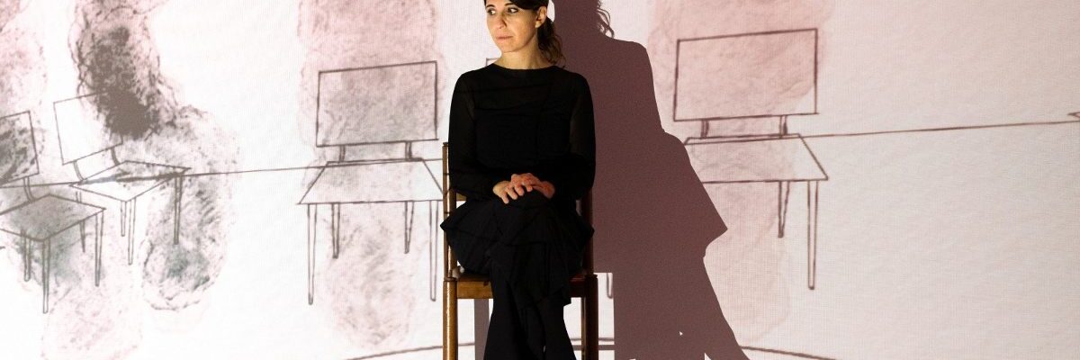 Una foto di scena delle spettacolo psychodrama: una donna seduta su una sedia di fronte a una grande proiezione dove ci sone disegnate della altre sedie, dislocate a destra e sinistra della donna