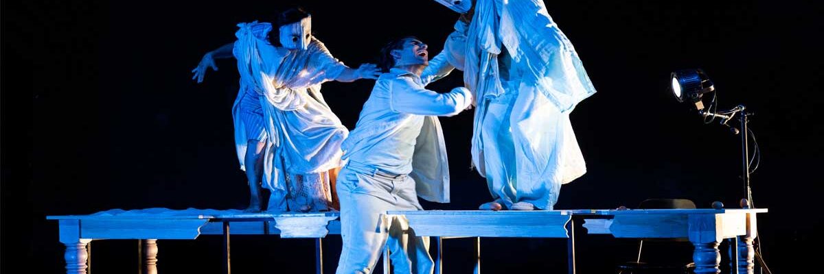 Scena dello spettacolo teatrale “Arrusi” con tre attori sul palco illuminati da luci drammatiche, in un’atmosfera intensa che richiama il tema della discriminazione e dei diritti negati.