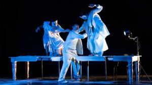 Scena dello spettacolo teatrale “Arrusi” con tre attori sul palco illuminati da luci drammatiche, in un’atmosfera intensa che richiama il tema della discriminazione e dei diritti negati.