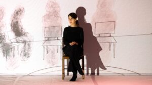 Una foto di scena delle spettacolo psychodrama: una donna seduta su una sedia di fronte a una grande proiezione dove ci sone disegnate della altre sedie, dislocate a destra e sinistra della donna