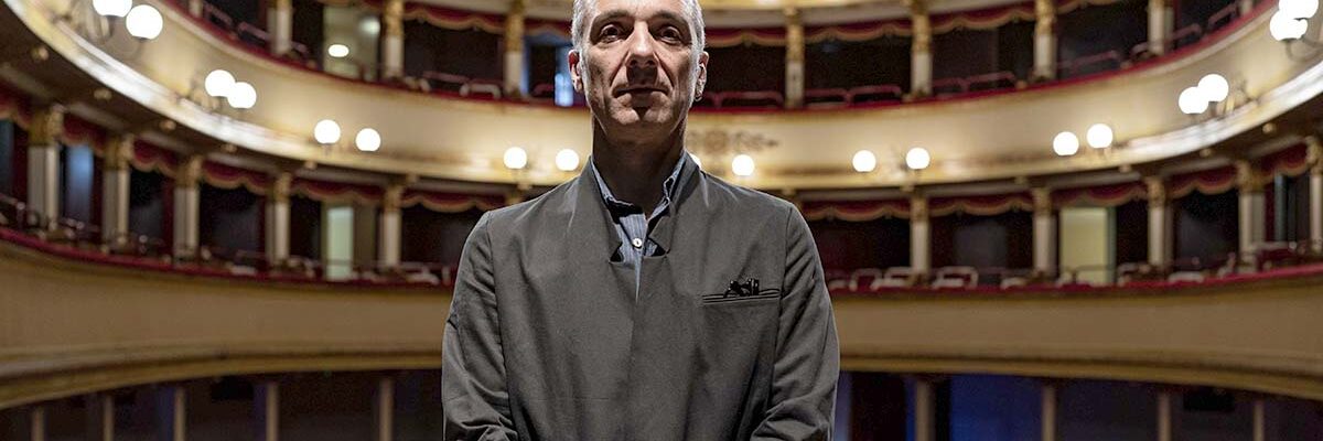 In questa foto, un uomo è in posa in piedi, dietro di lui si vede la platea di un grande teatro. L'uomo è Mauro Pescio, autore e attore dello spettacolo Io ero il Milanese.