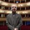 In questa foto, un uomo è in posa in piedi, dietro di lui si vede la platea di un grande teatro. L'uomo è Mauro Pescio, autore e attore dello spettacolo Io ero il Milanese.